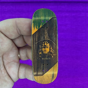 Peut inclure: Une planche à doigts en bois avec un motif de sorcier. Le dessus est vert et orange, avec une section centrale marron foncé représentant un sorcier et le texte "J. workers".