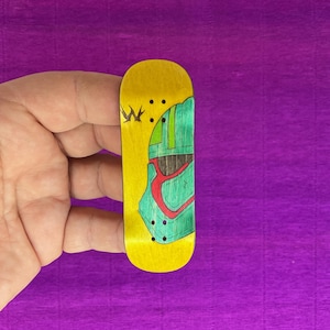 May include: A yellow fingerboard with a green and red graphic design. The miniature skateboard has black dots and a stylized letter "W" on the top. The background is a purple surface.