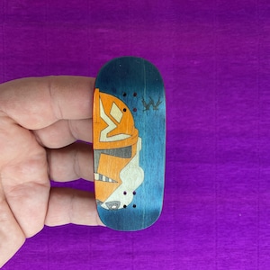 May include: A blue and orange fingerboard with a graphic design. The design features a stylized image of a helmet and a symbol. The fingerboard has a blue background and is made of wood.