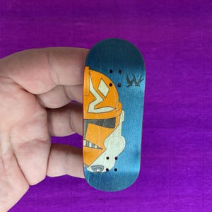 May include: A blue and orange fingerboard with a graphic design of a helmet. The fingerboard has a blue background with an orange helmet design on the left side and a small graphic on the right side.