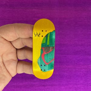May include: A yellow fingerboard with a green and red graphic design. The design features a helmet-like image. The fingerboard is held in a hand against a purple background. The fingerboard has a small logo on the top left.