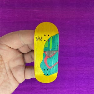 May include: A yellow fingerboard with a green and red graphic design. The fingerboard has a yellow base with a crown logo and three black dots. The graphic design features a helmet-like shape with green, red, and blue accents. The fingerboard is held in a hand.