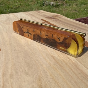 Puede incluir: Un soporte para incienso rectangular hecho de madera pulida y resina amarilla translúcida. La sección de madera presenta vetas de color marrón oscuro y símbolos negros grabados, incluida una figura de ave. Una varilla de latón descansa encima.
