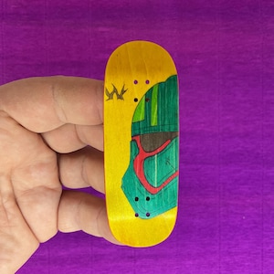 May include: A yellow fingerboard with a green and red graphic design. The design features a helmet-like shape on the top of the board. The fingerboard is held in a hand against a purple background.