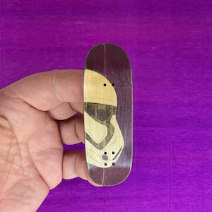 May include: A wooden fingerboard with a Stormtrooper helmet design. The top half of the board is light wood, while the bottom half is a dark brown. The helmet design is etched into the wood, with a detailed outline.