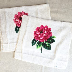 May include: Two white hand towels with a vibrant pink and red floral design. The flowers have green leaves and stems. The towels are made of a soft, absorbent material.