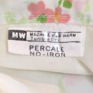 May include: Close-up of a white fabric with a floral pattern in pink, blue, and green. A label with the text "PERCALE NO-IRON" is visible. The label also includes washing instructions.