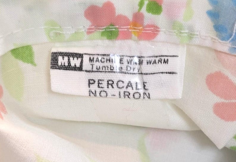May include: Close-up of a white fabric with a floral pattern in pink, blue, and green. A label with the text "PERCALE NO-IRON" is visible. The label also includes washing instructions.