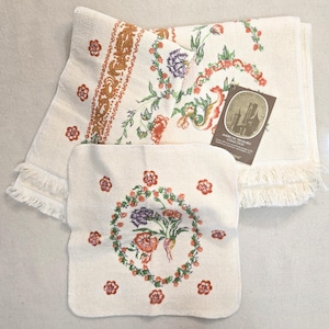May include: Off-white floral towel set featuring a large bath towel, a hand towel, and a washcloth. The towels have a floral design in shades of red, orange, green, and purple. The bath towel has a fringed edge and a decorative border.