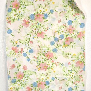 May include: A white fabric with a floral pattern featuring pink, blue, yellow, and white flowers with green leaves. The fabric is hanging from a clear plastic hanger.