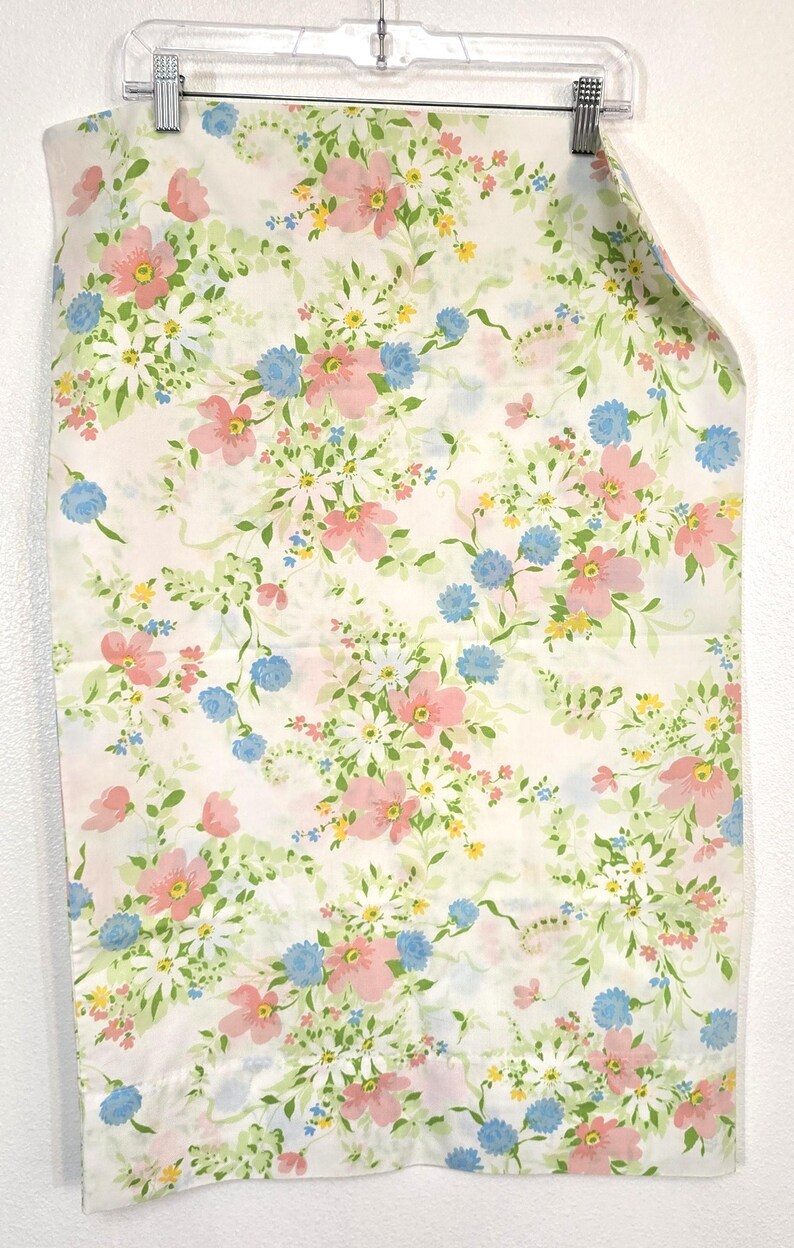 May include: A white fabric with a floral pattern featuring pink, blue, yellow, and white flowers with green leaves. The fabric is hanging from a clear plastic hanger.