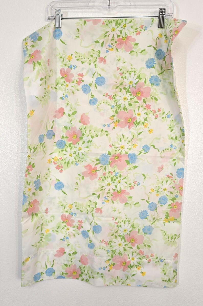 May include: A white fabric with a floral pattern featuring pink, blue, and yellow flowers, along with green leaves. The fabric is hanging from a clear plastic hanger.
