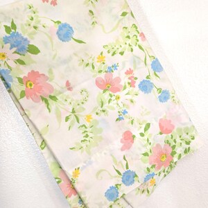 May include: A folded white sheet with a floral pattern. The pattern features pink, blue, and yellow flowers with green leaves and vines. The sheet is hanging from a metal hanger.