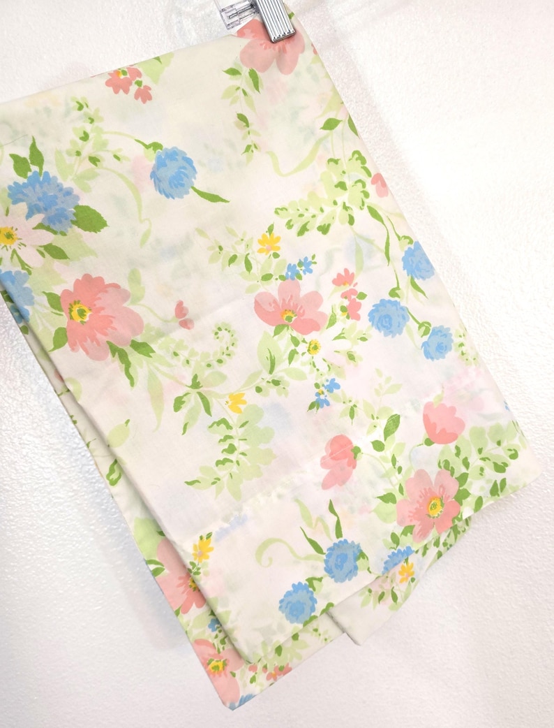 May include: A folded white sheet with a floral pattern. The pattern features pink, blue, and yellow flowers with green leaves and vines. The sheet is hanging from a metal hanger.