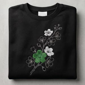May include: Black crewneck sweatshirt featuring an embroidered floral design. The design includes white and green blossoms with gray stems and outlines. The sweatshirt has a ribbed collar and a blank white tag.