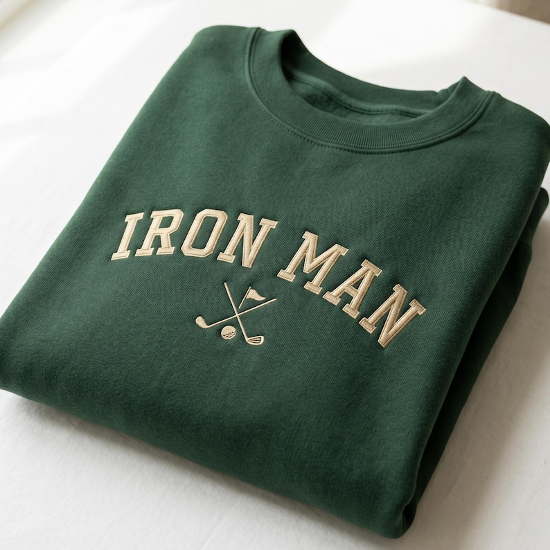 May include: A folded, dark green sweatshirt with the embroidered text "IRON MAN" in a tan, arched font. Below the text is a golf club and flag design. The sweatshirt is made of a soft material.
