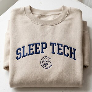 Könnte beinhalten: Beiges Sweatshirt mit marineblau gesticktem Text "SLEEP TECH" in einem Bogen, über einer marineblauen Grafik eines schlafenden Fuchses in einem Mond mit Sternen. Das Sweatshirt ist auf einer weißen Oberfläche gefaltet.