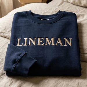 Puede incluir: Sudadera azul marino con la palabra "LINEMAN" bordada en color beige claro en el pecho. La sudadera está doblada cuidadosamente sobre una cama, mostrando el cuello redondo y los puños acanalados.