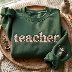 May include: A dark green sweatshirt with the word "teacher" embroidered in floral letters. The sleeve cuff features the name "Rebecca" and a small flower. The sweatshirt is folded on a wooden tray.