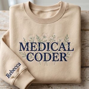 May include: A beige crewneck sweatshirt featuring dark blue embroidered text that reads MEDICAL CODER, surrounded by delicate floral line art in green and white. The sleeve is embroidered with the name Rebecca in dark blue thread.