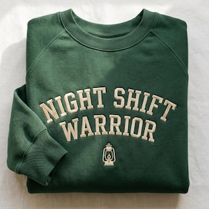 Puede incluir: Sudadera verde oscuro con la inscripción bordada "NIGHT SHIFT WARRIOR" en beige claro. Debajo, un gráfico de una linterna. Cuello redondo y mangas largas. Prenda informal.