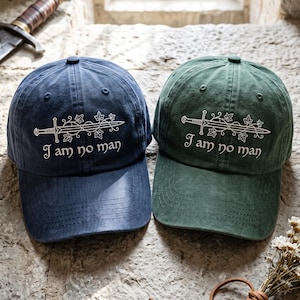 May include: Two baseball caps, one navy blue and one forest green, each featuring an embroidered white design of a sword entwined with ivy vines and the text "I am no man" in a stylized font. Both caps have a structured crown and curved brim.