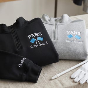 Puede incluir: Una sudadera negra y una gris con cremallera de un cuarto, cada una con texto bordado que dice PAHS Color Guard sobre dos banderas azul claro cruzadas. La sudadera negra incluye la palabra Custom bordada en blanco en el puño de la manga.