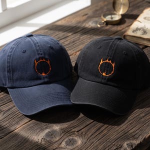 May include: Two baseball caps, one in navy blue and one in black, each featuring an embroidered orange circular flame design on the front panel. Both caps have a structured crown, a curved brim, and stitched eyelets for ventilation.