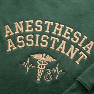May include: A dark green sweatshirt with the embroidered text "ANESTHESIA ASSISTANT" in a tan, collegiate-style font. Below the text is a medical symbol with a heartbeat line. The sweatshirt has a ribbed cuff.
