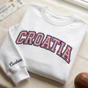 May include: A white crewneck sweatshirt featuring the word CROATIA embroidered in a bold, arched collegiate font with red and navy blue outlines. The left sleeve cuff area displays the word Custom embroidered in navy blue script lettering.