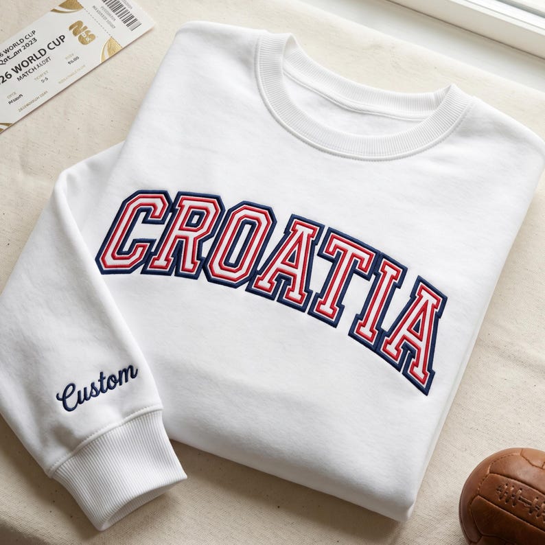May include: A white crewneck sweatshirt featuring the word CROATIA embroidered in a bold, arched collegiate font with red and navy blue outlines. The left sleeve cuff area displays the word Custom embroidered in navy blue script lettering.