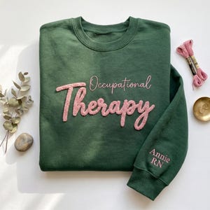 May include: A dark green crewneck sweatshirt featuring the text Occupational Therapy in pink textured chenille lettering across the chest. The sleeve includes the embroidered name Annie RN in pink thread. The garment has a ribbed collar and cuffs.