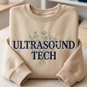 Könnte beinhalten: Ein beigefarbenes Sweatshirt mit Rundhalsausschnitt und dem dunkelblau gestickten Text "ULTRASOUND TECH" mittig auf der Brust. Über und hinter dem Text befindet sich ein gesticktes Blumenstrauß-Design in Salbeigrün, Weiß und Hellblau.