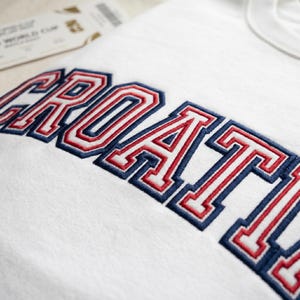 May include: A white sweatshirt featuring the word CROATIA embroidered in a collegiate font. The lettering is outlined in dark blue with a red inner border, creating a layered effect. The fabric is a plain white knit material with a crew neckline.