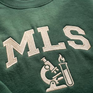Puede incluir: Sudadera verde con texto bordado "MLS" en crema, en una fuente de estilo universitario. Debajo del texto hay un gráfico bordado en crema de un microscopio y un tubo de ensayo con una llama. La sudadera es de material suave.
