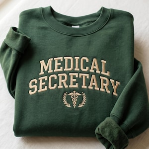 Könnte beinhalten: Dunkelgrünes Sweatshirt mit dem gestickten Schriftzug "MEDICAL SECRETARY" in hellem Beige. Darunter befindet sich ein medizinisches Symbol. Das Sweatshirt hat einen Rundhalsausschnitt und gerippte Bündchen.