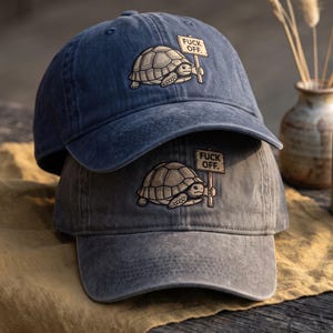 May include: Two baseball caps, one dark blue and one grey, each featuring an embroidered graphic of a turtle holding a sign that reads FUCK OFF. The caps have a distressed, washed-out fabric texture and a curved brim with visible stitching details.