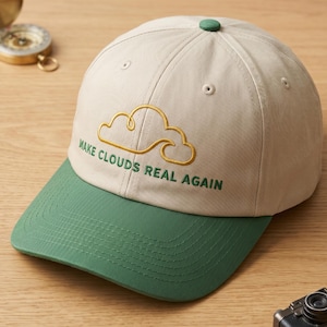 May include: A beige baseball cap with a green brim and a green button on top. The front features an embroidered gold outline of a cloud above the green text that reads, "MAKE CLOUDS REAL AGAIN." The cap has stitched eyelets on the crown panels.