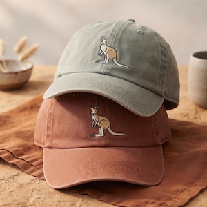 May include: A sage green baseball cap stacked on top of a terracotta baseball cap. Both caps feature an embroidered tan kangaroo graphic centered on the front panel. The caps have a curved brim, stitched eyelets, and a structured six-panel design.