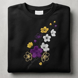 May include: Black crewneck sweatshirt featuring an embroidered floral design. The design includes yellow, purple, and white flowers on a dark branch. The sweatshirt is folded neatly on a neutral surface.