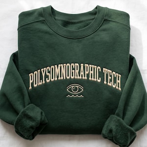 Könnte beinhalten: Dunkelgrünes Sweatshirt mit cremefarbenem, bogenförmigem Text "POLYSOMNOGRAPHIC TECH". Darunter ein Augensymbol über Wellenlinien. Die Ärmel sind umgeschlagen, das Sweatshirt ist gefaltet.