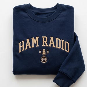 May include: A folded navy blue sweatshirt featuring the embroidered words "HAM RADIO" in tan. Below the text is a tan microphone and globe design. The sweatshirt is neatly folded.