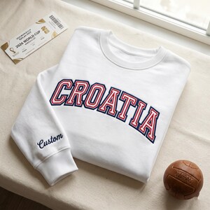 May include: A white crewneck sweatshirt featuring the word CROATIA embroidered in a red and navy blue collegiate font across the chest. The word Custom is embroidered in navy blue on the sleeve cuff. A 2026 World Cup ticket and a brown leather ball.