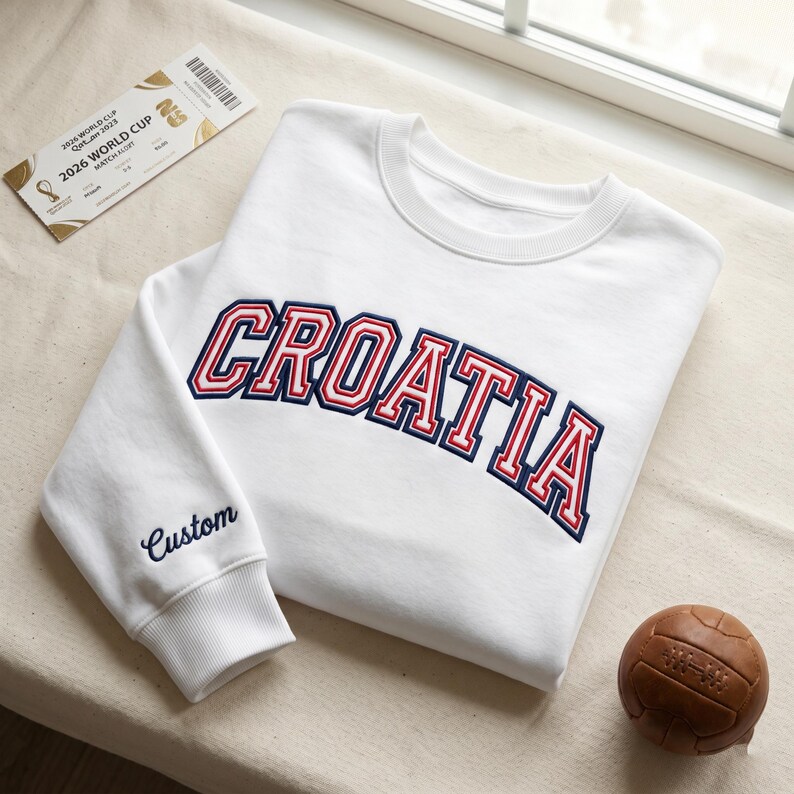 May include: A white crewneck sweatshirt featuring the word CROATIA embroidered in a red and navy blue collegiate font across the chest. The word Custom is embroidered in navy blue on the sleeve cuff. A 2026 World Cup ticket and a brown leather ball.