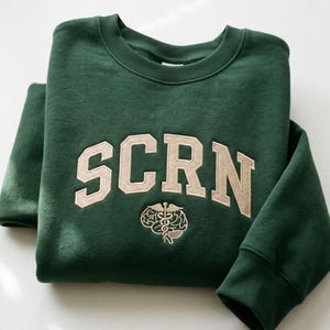 Può includere: Una felpa verde scuro piegata con la scritta "SCRN" in beige, in stile collegiale, sul petto. Sotto la scritta c'è una grafica di un cervello beige con un simbolo di caduceo. La felpa ha un girocollo e polsini a coste.