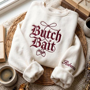 Puede incluir: Una sudadera de cuello redondo color crema con bordado rojo oscuro en el pecho que dice "Butch Bait" en una fuente gótica con un lazo y dos fresas. La manga izquierda está bordada con el nombre "Rebecca" en letra cursiva de color rojo.