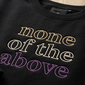 May include: Black sweatshirt with the embroidered phrase "none of the above" in a gradient of colors. The top text is gold, the middle text is white, and the bottom text is purple. The sweatshirt has a ribbed collar.