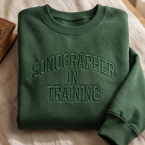 Puede incluir: Una sudadera verde oscuro con el texto bordado "SONOGRAPHER IN TRAINING" en hilo verde a juego. La sudadera tiene cuello redondo y mangas largas. La tela parece suave y cómoda.