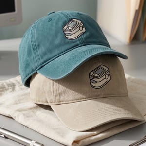 May include: A teal baseball cap stacked on top of a beige baseball cap. Each cap features an embroidered graphic of a pulse oximeter on the front panel. The caps have a structured crown, a curved brim, and a classic six-panel design with stitching.