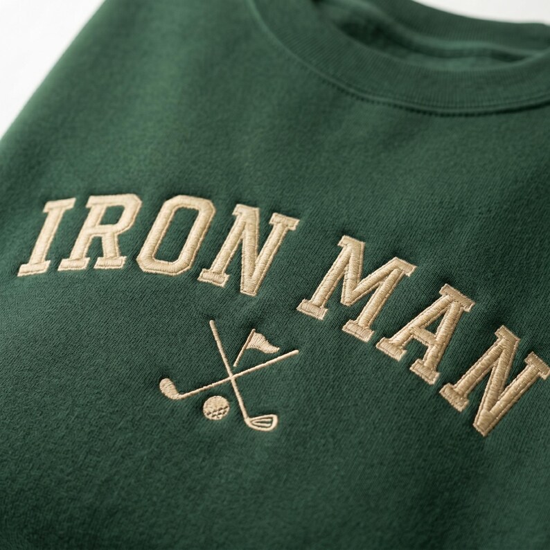 May include: A dark green sweatshirt featuring the embroidered words "IRON MAN" in a light tan hue. Below the text is a golf-themed emblem with crossed golf clubs, a golf ball, and a flag.